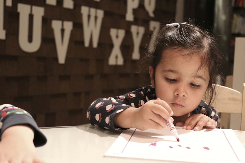 Young girl drawing at a white table in front of a shingled wall with large cutout letters mounted on it